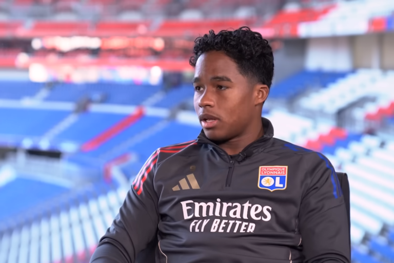 O jogador brasileiro Endrick durante uma entrevista vestindo agasalho de treino cinza do clube francês Olympique Lyonnais em um estádio de futebol.