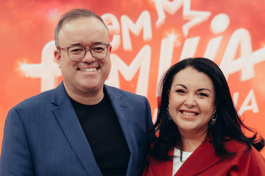 Everaldo Marques, de óculos e paletó azul, sorrindo ao lado de sua esposa Iane Marques, que veste um casaco vermelho, em frente a um painel de fundo laranja com texto branco em um evento.