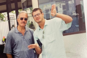 Fábio Assunção jovem, usando óculos de grau e camisa clara, acena com a mão esquerda levantada ao lado de seu pai, Ivo Assunção Pinto, que veste camisa polo cinza, óculos de sol e tem bigode. O registro antigo mostra os dois em um ambiente externo iluminado.