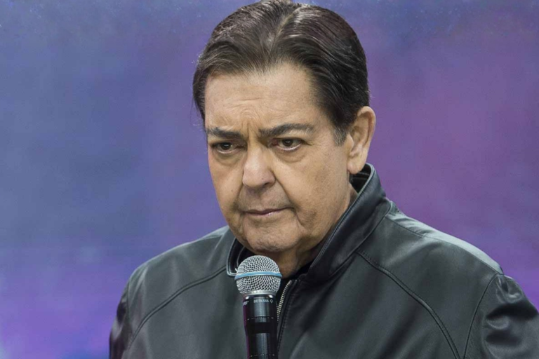 O apresentador Fausto Silva, o Faustão, com expressão séria, vestindo uma jaqueta de couro preta e segurando um microfone próximo ao rosto, contra um fundo desfocado em tons de roxo.