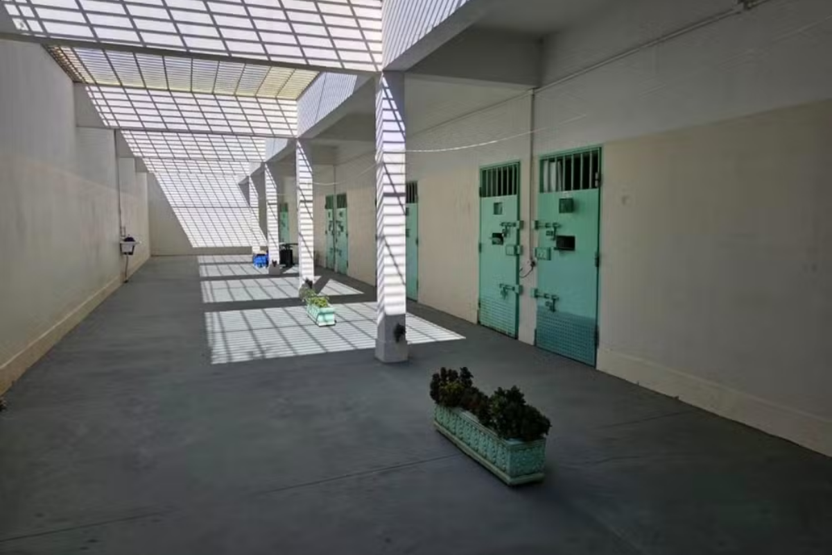 Corredor externo de uma unidade prisional com piso de concreto, colunas brancas e portas metálicas verdes com grades e pequenas aberturas, sob uma cobertura metálica vazada que projeta sombras geométricas no chão, com vasos de plantas no centro.