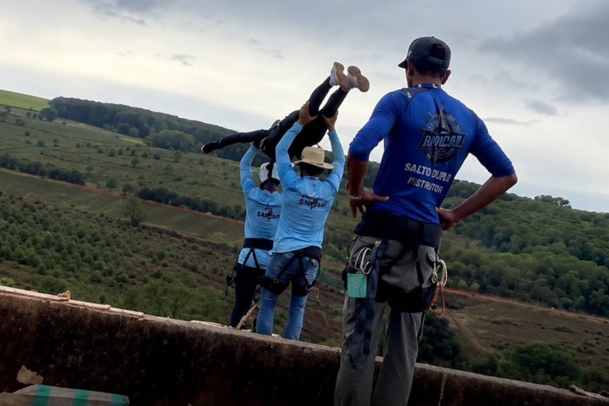 Instrutores de salto preparam pessoa para prática de rope jump em local de altura elevada com paisagem montanhosa ao fundo.