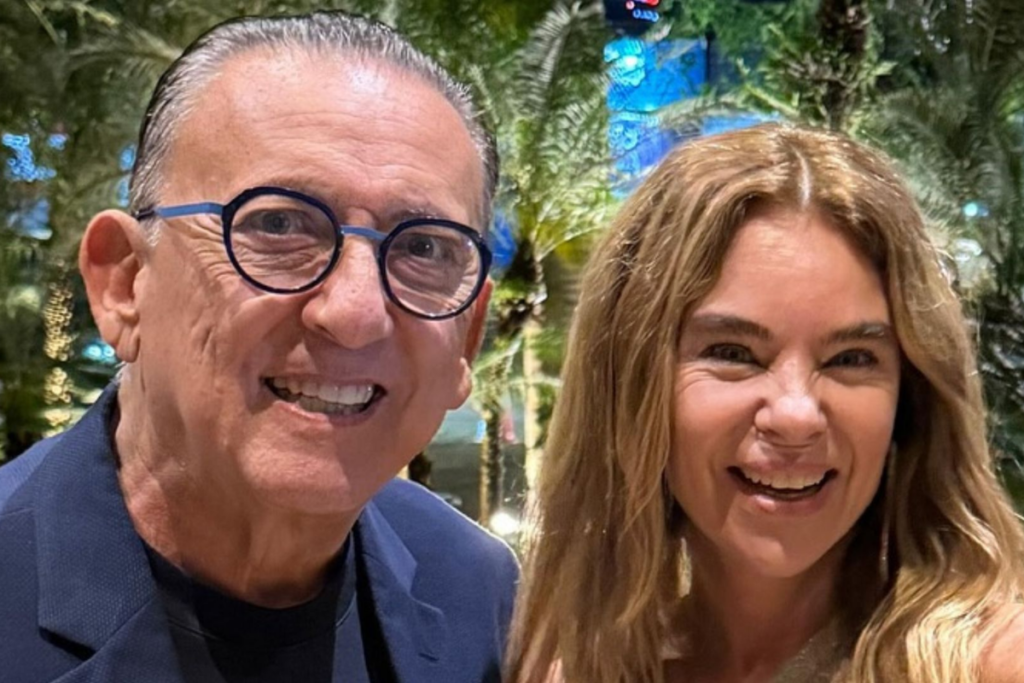 Galvão Bueno, usando óculos de armação azul e paletó, posa sorrindo ao lado de sua esposa, Desirée Soares, que tem cabelos longos e loiros, em ambiente noturno com iluminação externa.