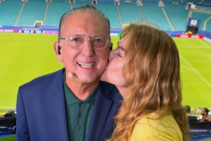 Galvão Bueno sorri para a câmera usando terno azul, camisa verde, óculos e fone de ouvido, enquanto recebe um beijo na bochecha de sua esposa, Desirée Soares, que tem cabelos loiros ondulados. Ao fundo, vê-se o gramado verde e as arquibancadas vazias de um estádio de futebol.
