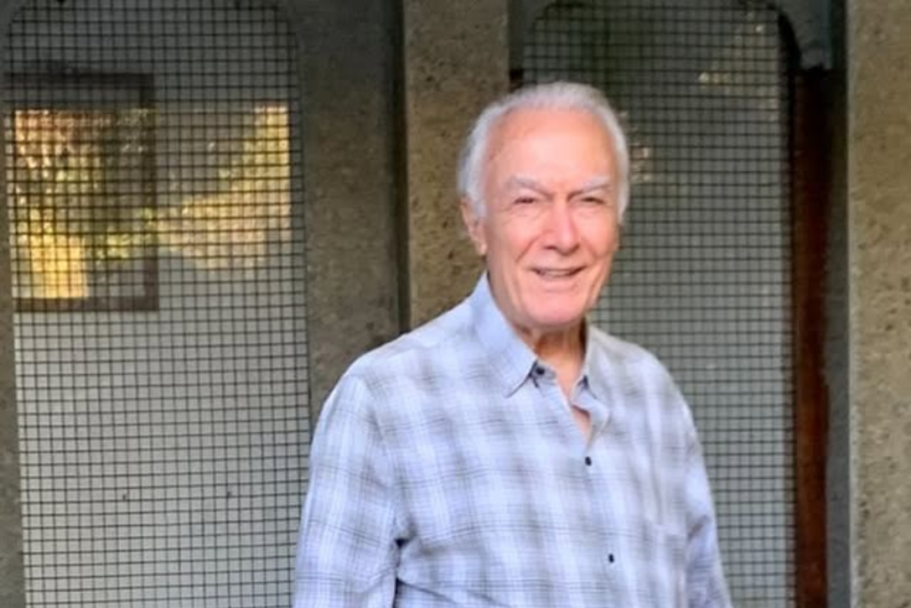 Ivo Assunção Pinto, um homem idoso de cabelos brancos e sorridente, veste uma camisa social de manga longa com estampa xadrez em tons de cinza e branco. Ele está em um ambiente com fundo cinza estruturado e detalhes em malha quadriculada.