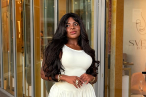 Jojo Todynho sorri de forma confiante, vestindo um vestido branco justo e com cabelos longos e escuros soltos, posando na entrada de uma loja sofisticada com detalhes dourados. Ela tem acessórios dourados e um anel grande,
