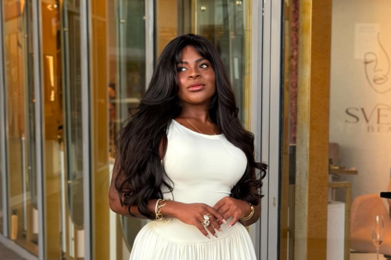 Jojo Todynho sorri de forma confiante, vestindo um vestido branco justo e com cabelos longos e escuros soltos, posando na entrada de uma loja sofisticada com detalhes dourados. Ela tem acessórios dourados e um anel grande,