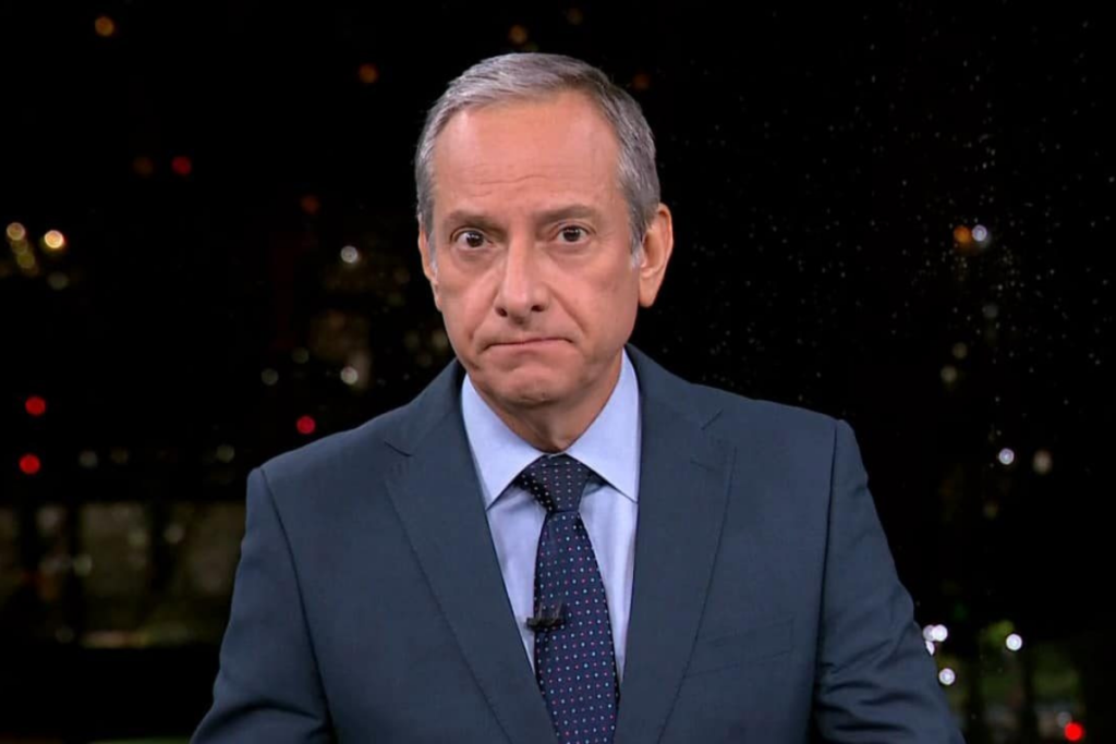 O jornalista José Roberto Burnier vestindo terno e gravata, olhando diretamente para a câmera contra um fundo escuro com luzes desfocadas.