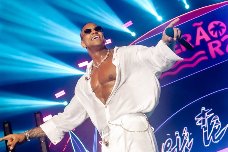 O cantor Léo Santana sorridente durante show, usando óculos de sol escuros, camisa branca aberta exibindo o peitoral, calça branca com detalhe em corrente e colar de corrente prateada. Ele segura um microfone e aponta para o alto em um palco com luzes azuis ao fundo e painel de LED.