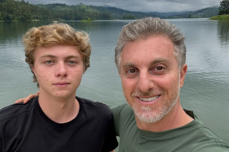 Uma foto de close-up do apresentador de TV brasileiro Luciano Huck sorrindo e com o braço em volta do pescoço de seu filho adolescente, Benício, em uma paisagem externa. Luciano Huck, à direita, tem cabelos grisalhos curtos e uma barba bem cuidada. Ele está vestindo uma camiseta verde-oliva. Benício Huck, à esquerda, tem cabelos loiros encaracolados e está vestindo uma camiseta preta. Os dois estão de pé em frente a um grande lago azul com colinas verdes e frondosas e um céu nublado ao fundo. A expressão de Benício é séria, enquanto Luciano está sorrindo calorosamente para a câmera.