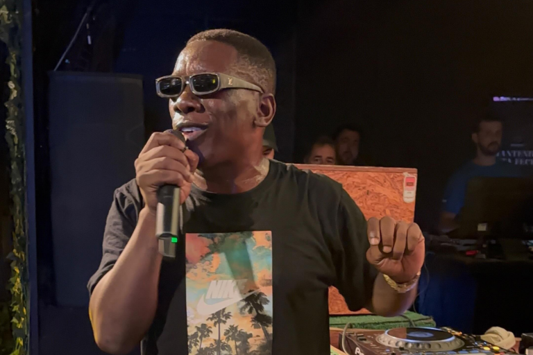 MC Gorila canta em microfone durante show usando camiseta preta estampada e óculos escuros de grife em ambiente noturno.