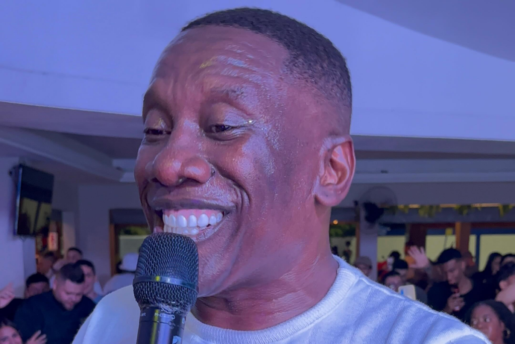MC Gorila sorrindo enquanto fala ao microfone em um evento com público ao fundo.