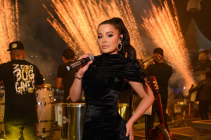 Cantora Maraisa posa segurando um microfone no palco durante um show à noite. Ela veste um vestido preto de veludo, tem os cabelos presos em um rabo de cavalo alto e usa brincos grandes dourados. Ao fundo, integrantes da banda tocam instrumentos e há uma queima de fogos de artifício com faíscas douradas iluminando o cenário.