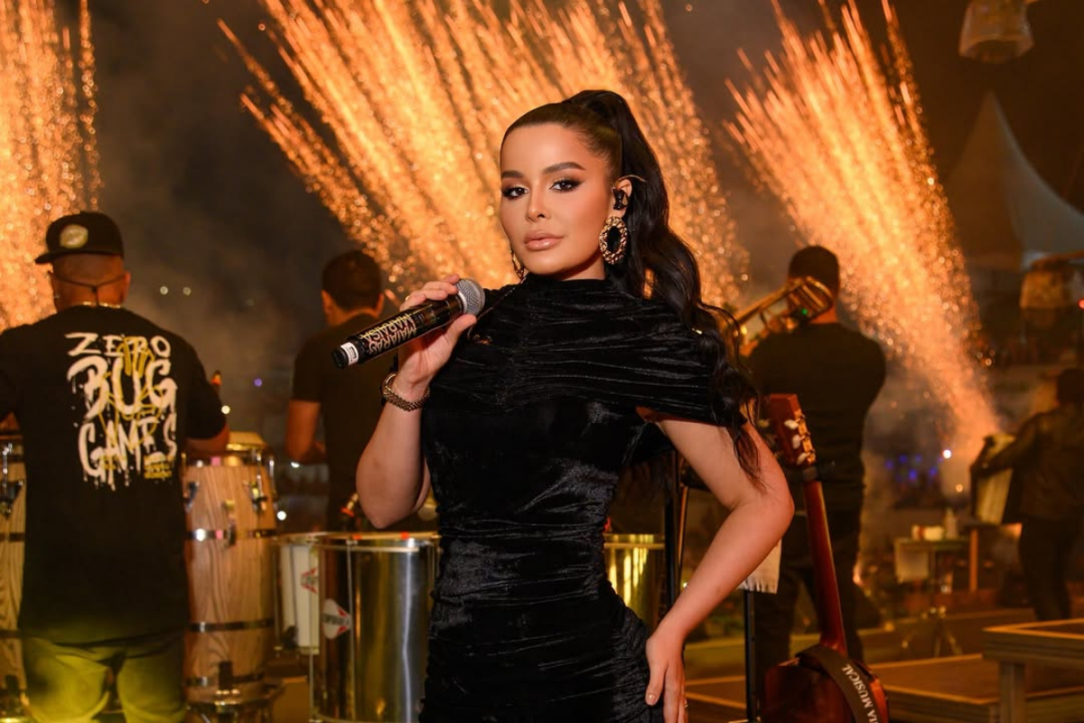 Cantora Maraisa posa segurando um microfone no palco durante um show à noite. Ela veste um vestido preto de veludo, tem os cabelos presos em um rabo de cavalo alto e usa brincos grandes dourados. Ao fundo, integrantes da banda tocam instrumentos e há uma queima de fogos de artifício com faíscas douradas iluminando o cenário.