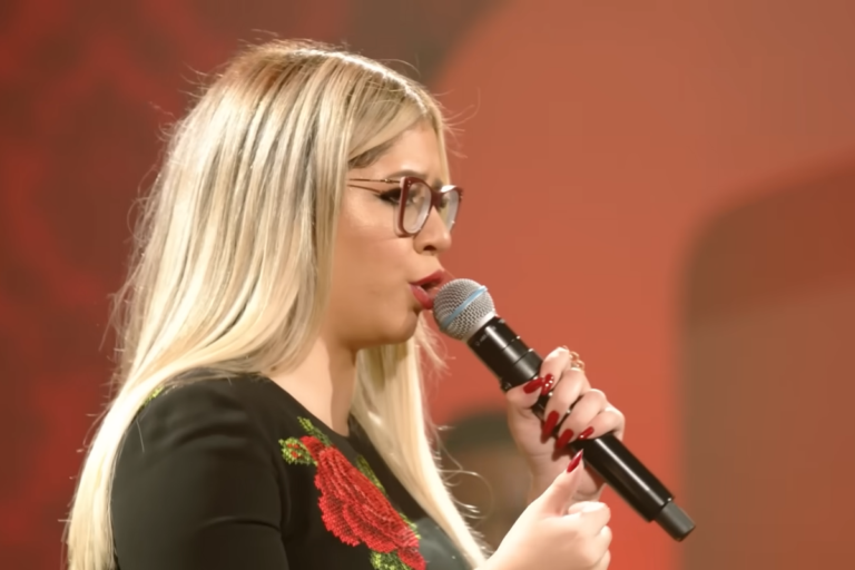 Marília Mendonça com cabelos loiros e óculos de grau, cantando ao microfone com expressão intensa durante apresentação musical.