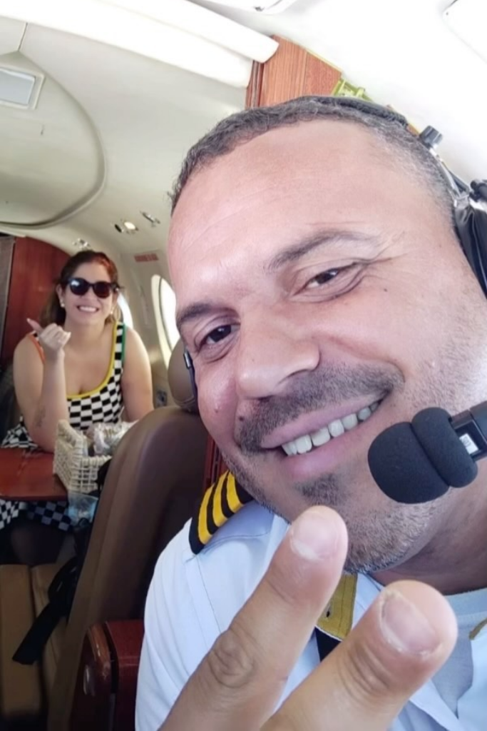Marília Mendonça sorrindo dentro de uma aeronave enquanto o piloto faz sinal de paz com a mão para a câmera em primeiro plano.
