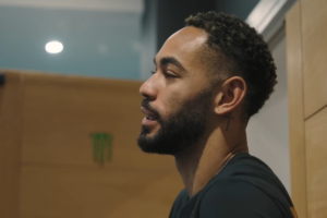 Jogador de futebol Matheus Cunha visto de perfil, com barba curta, tatuagem de cruz atrás da orelha e vestindo camisa escura em ambiente interno.