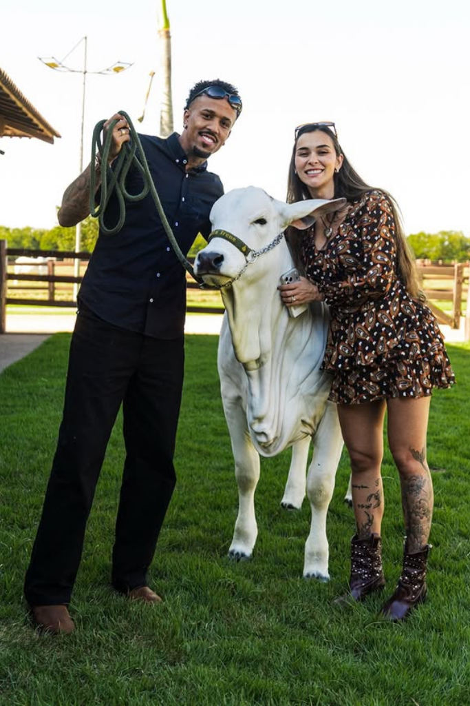 Eder Militão e Tainá Castro sorriem em pé em uma área de gramado ao ar livre ao lado de um bezerro branco de raça.