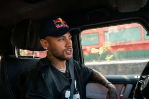 Jogador de futebol Neymar de perfil, sentado no banco do motorista de um carro, usando boné azul-marinho da Red Bull, camiseta preta da Puma, colar de brilhantes e brincos. Ele tem barba por fazer, tatuagens no braço e no pescoço, e olha para a direita.
