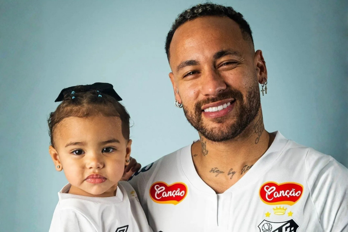 O jogador de futebol Neymar sorrindo e segurando no colo sua filha bebê, Mavie. Ambos vestem camisas brancas oficiais do Santos Futebol Clube, com o patrocínio da marca Canção em destaque. O fundo da imagem é um tom de azul claro e liso.