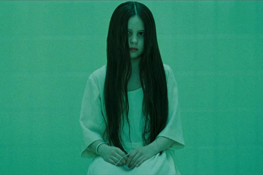 Atriz Daveigh Chase interpretando a personagem Samara Morgan em uma cena do filme de terror O Chamado, sentada e olhando diretamente com os longos cabelos pretos cobrindo parcialmente o rosto em um ambiente monocromático verde.