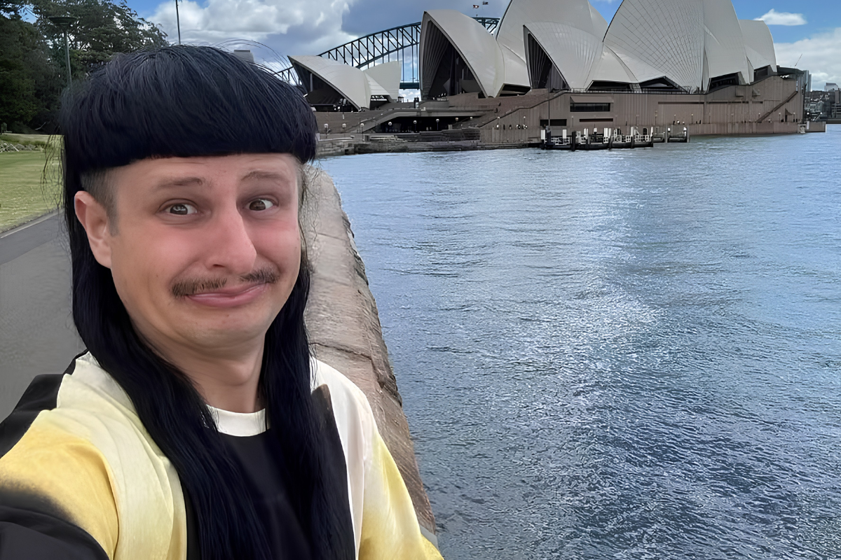 O cantor Oliver Tree sorri para uma selfie em primeiro plano, exibindo seu característico corte de cabelo estilo tigela com mechas longas e bigode. Ao fundo, destaca-se a imponente arquitetura da Ópera de Sydney e a Harbour Bridge sob um céu parcialmente nublado na Austrália.