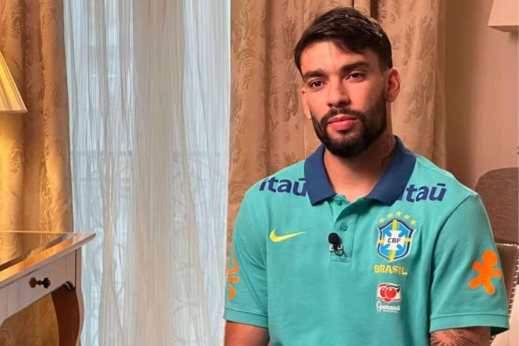 O jogador de futebol Lucas Paquetá, com barba e cabelo escuro, posa para uma foto em ambiente interno. Ele veste uma camisa polo azul-turquesa da Seleção Brasileira, com os patrocinadores Itaú e Guaraná visíveis, além do escudo da CBF.