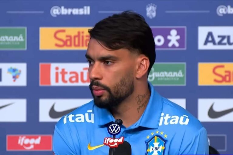 Lucas Paquetá em entrevista coletiva, vestindo a camisa azul de treino da Seleção Brasileira com logotipos de patrocinadores ao fundo.