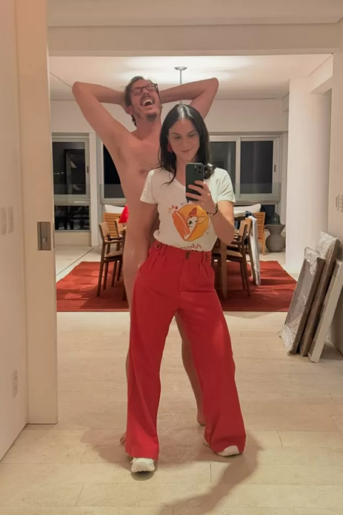 Priscila Castello Branco tira uma foto no espelho usando camiseta branca estampada e calça vermelha, enquanto Fábio Porchat aparece ao fundo, rindo com as mãos na cabeça, simulando estar nu atrás dela.