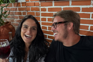 Priscila Castello Branco sorri alegremente segurando uma taça com bebida escura, enquanto o noivo Fábio Porchat, usando óculos de grau, olha para ela e ri, em um ambiente com parede de tijolos ao fundo.