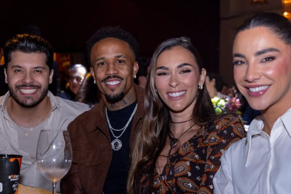 Murilo Huff, Eder Militão, Tainá Castro e Gabriela Versiani sorriem sentados juntos ao redor de uma mesa redonda durante um evento social noturno em ambiente fechado.