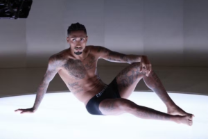 O jogador de futebol Raphinha posa sentado sobre uma superfície iluminada, vestindo apenas roupas íntimas pretas, exibindo suas diversas tatuagens no peito, braços e pernas.