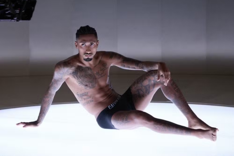 O jogador de futebol Raphinha posa sentado sobre uma superfície iluminada, vestindo apenas roupas íntimas pretas, exibindo suas diversas tatuagens no peito, braços e pernas.