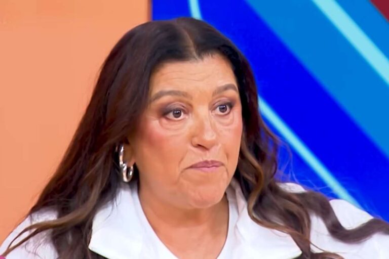 Primeiro plano da apresentadora Regina Casé com cabelos longos e escuros, usando uma blusa branca e brincos de argola, olhando para a frente com expressão séria.
