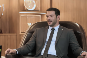 Presidente da CBF, Samir Xaud, sentado em uma poltrona de couro marrom em um cenário com troféus ao fundo, vestindo terno cinza, camisa social clara e gravata.