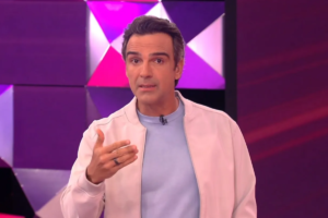 Apresentador Tadeu Schmidt, homem de pele clara e cabelos escuros penteados para trás, vestindo camiseta azul-clara sob uma jaqueta off-white. Ele está com a mão direita levantada fazendo um gesto, posicionado em frente a um cenário com formas geométricas em tons de roxo e rosa.