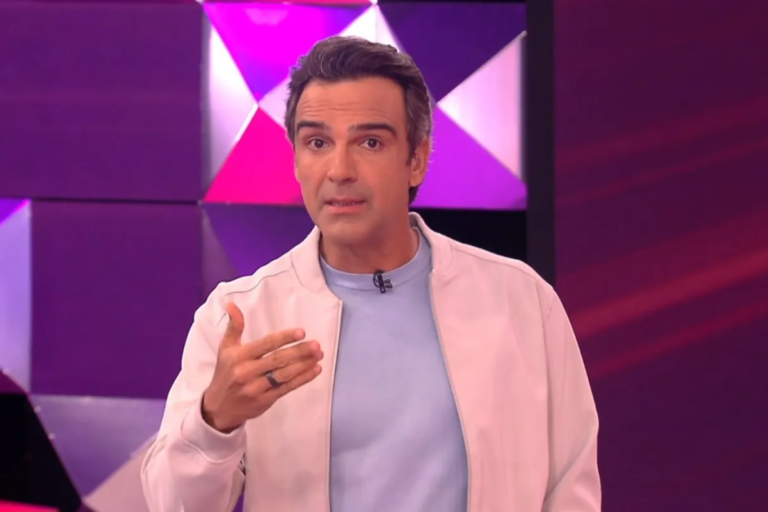 Apresentador Tadeu Schmidt, homem de pele clara e cabelos escuros penteados para trás, vestindo camiseta azul-clara sob uma jaqueta off-white. Ele está com a mão direita levantada fazendo um gesto, posicionado em frente a um cenário com formas geométricas em tons de roxo e rosa.