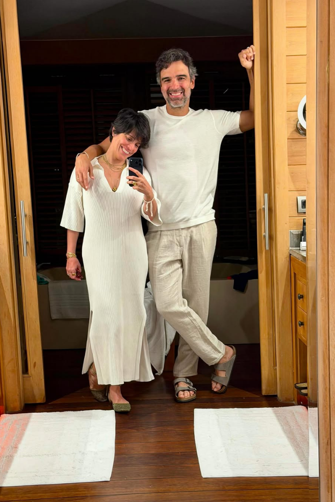 O apresentador Tadeu Schmidt e sua esposa, Ana Cristina Schmidt, posam sorridentes para uma foto no espelho em um ambiente com decoração em madeira. Tadeu está abraçando Ana Cristina pelos ombros, vestindo uma camiseta branca, calça de linho bege claro e sandálias. Ana Cristina segura o celular para tirar a foto, veste um vestido longo canelado na cor branca com fendas laterais, colares dourados e sapatilhas.
