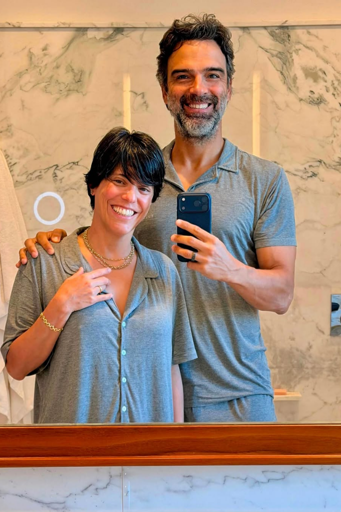 O apresentador Tadeu Schmidt e sua esposa, Ana Cristina Schmidt, sorriem em uma selfie tirada no espelho do banheiro. Ambos usam pijamas combinando na cor cinza-mescla. Tadeu segura o celular com uma das mãos e tem o outro braço sobre o ombro de Ana Cristina, que usa colares dourados e uma pulseira. Ao fundo, destaca-se uma parede de mármore claro com veios acinzentados e um espelho com detalhe circular iluminado.