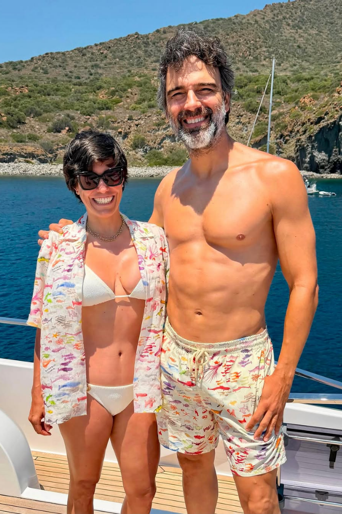 O jornalista e apresentador Tadeu Schmidt sorri ao lado de sua esposa, Ana Cristina Schmidt, a bordo de um barco durante o dia. Ele está sem camisa, vestindo uma bermuda de banho estampada. Ela usa óculos de sol escuros, biquíni branco e uma camisa de botões aberta com a mesma estampa da bermuda dele. Ao fundo, uma paisagem com águas azuis e morros com vegetação sob o céu claro.
