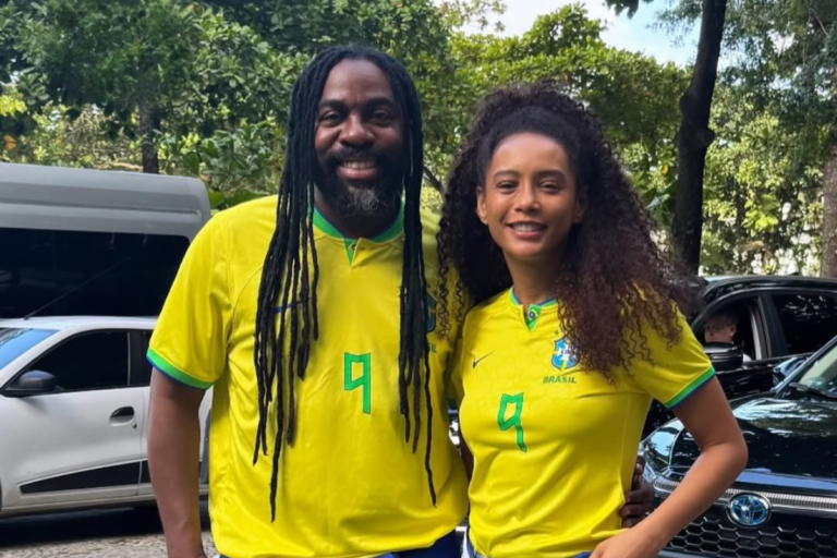 Lázaro Ramos, homem negro com dreadlocks longos, sorri ao lado de Taís Araújo, mulher negra com cabelos cacheados volumosos. Ambos vestem a camisa amarela da Seleção Brasileira de Futebol, com detalhes em verde e o número 9 na frente. Eles estão em um ambiente externo com árvores e carros ao fundo.