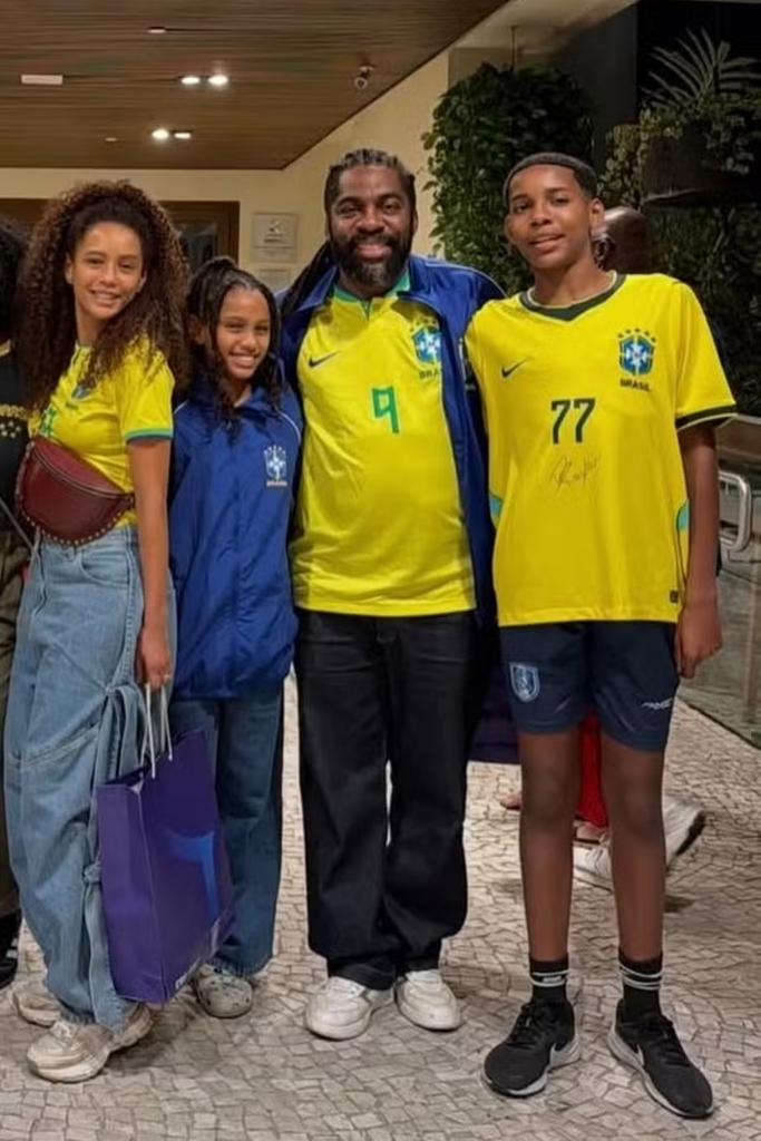 Taís Araújo, Lázaro Ramos e seus dois filhos, João Vicente e Maria Antônia, posam juntos sorrindo em um ambiente interno. Todos vestem roupas da Seleção Brasileira de Futebol: as mulheres e o jovem usam camisas amarelas, enquanto Lázaro veste uma camisa amarela sob um casaco azul. Eles estão de calças jeans e bermuda, e a filha segura uma sacola de compras azul.