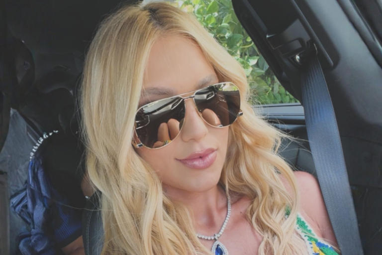 Virginia Fonseca com cabelos loiros ondulados, usando óculos de sol modelo aviador e colar com pingente grande, sentada no banco do passageiro de um carro.
