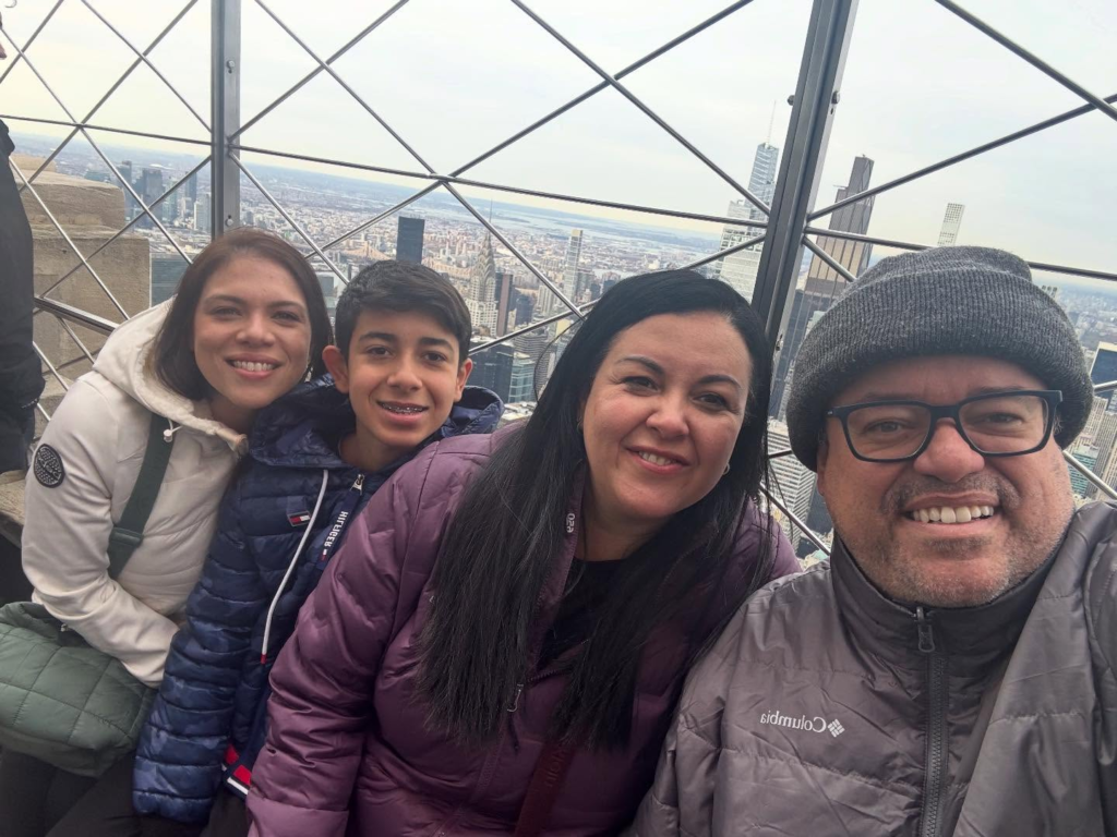 Everaldo Marques, sua esposa Iane e seus dois filhos sorrindo para uma selfie em um observatório de arranha-céu com vista panorâmica de Nova York ao fundo.