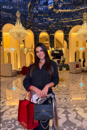 Tamires Fernandes Barcellos posa sorridente em um saguão de hotel luxuoso, segurando uma bolsa de luxo preta e uma sacola de compras vermelha.