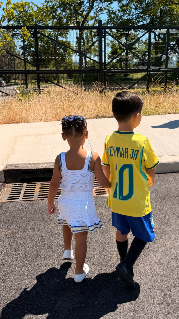 Mavie, filha de Neymar e Bruna Biancardi, de mãos dadas com um menino usando a camisa da seleção brasileira com o nome Neymar Jr, durante visita a um campo de treino.