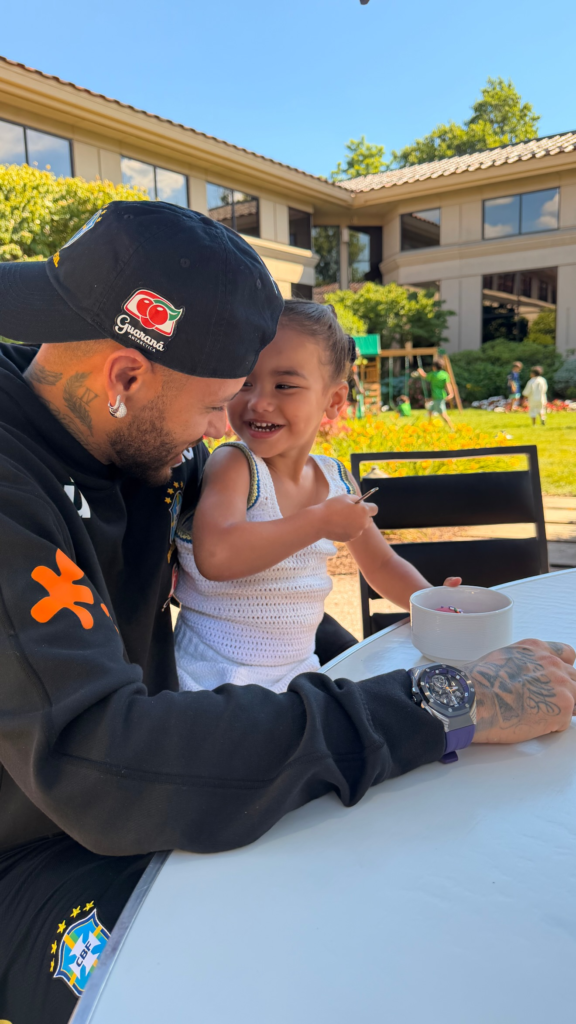 Neymar sorrindo enquanto interage com sua filha Mavie, que segura uma colher e senta-se à mesa em uma área externa durante um dia de treino.