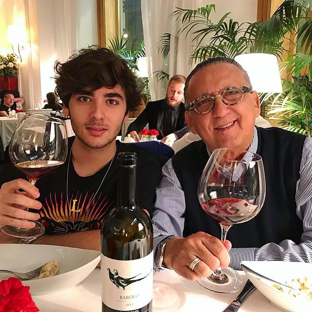 Galvão Bueno e seu filho Luca Bueno sentados à mesa de um restaurante, segurando taças de vinho tinto, com uma garrafa de vinho Barolo posicionada entre eles.
