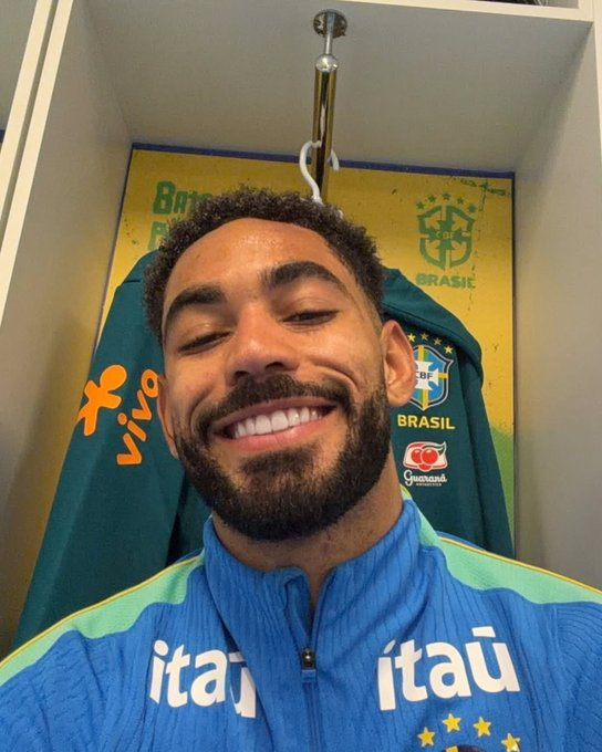 Jogador Matheus Cunha sorridente posando para selfie dentro do vestiário da Seleção Brasileira, vestindo casaco de treino azul com patrocínio do Itaú.