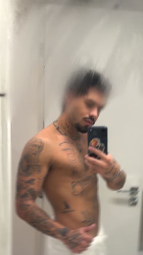 O cantor Zé Felipe, sem camisa e com o corpo tatuado, faz uma selfie em frente a um espelho de banheiro, segurando um smartphone. Ele está envolto em uma toalha branca e sorri levemente.